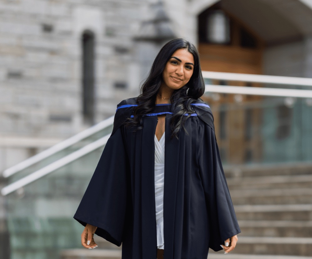 A graduate stands in a black graduation gown in front of a gray brick building.