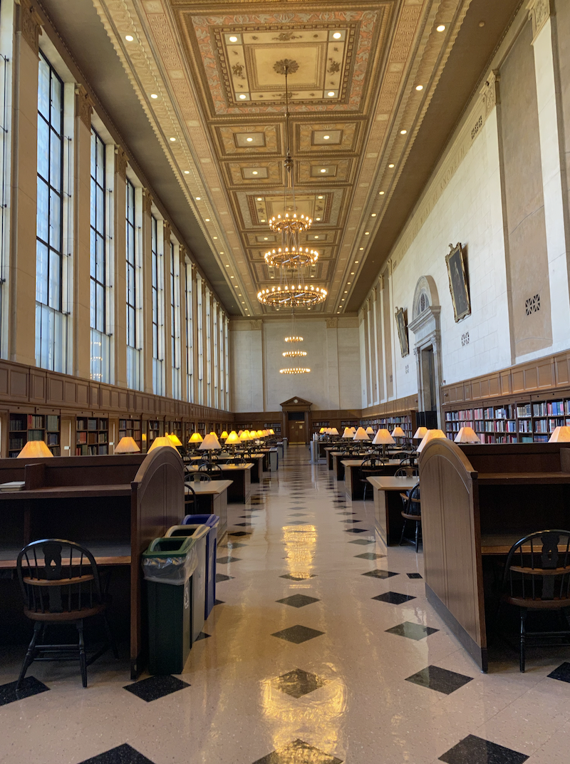 The main reading room of Butler Library at Columbia University. There are desks on either side of an aisle and a golden chandelier in the back.