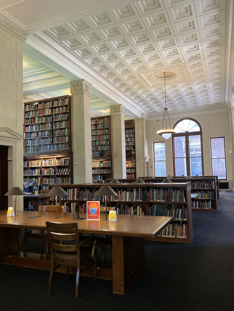 The inside of a library with shelves of books to the left and wooden tables in the middle.