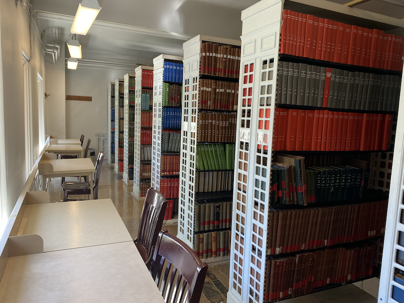 The inside of a library with stacks of books to the right and metal tables with chairs to the left.