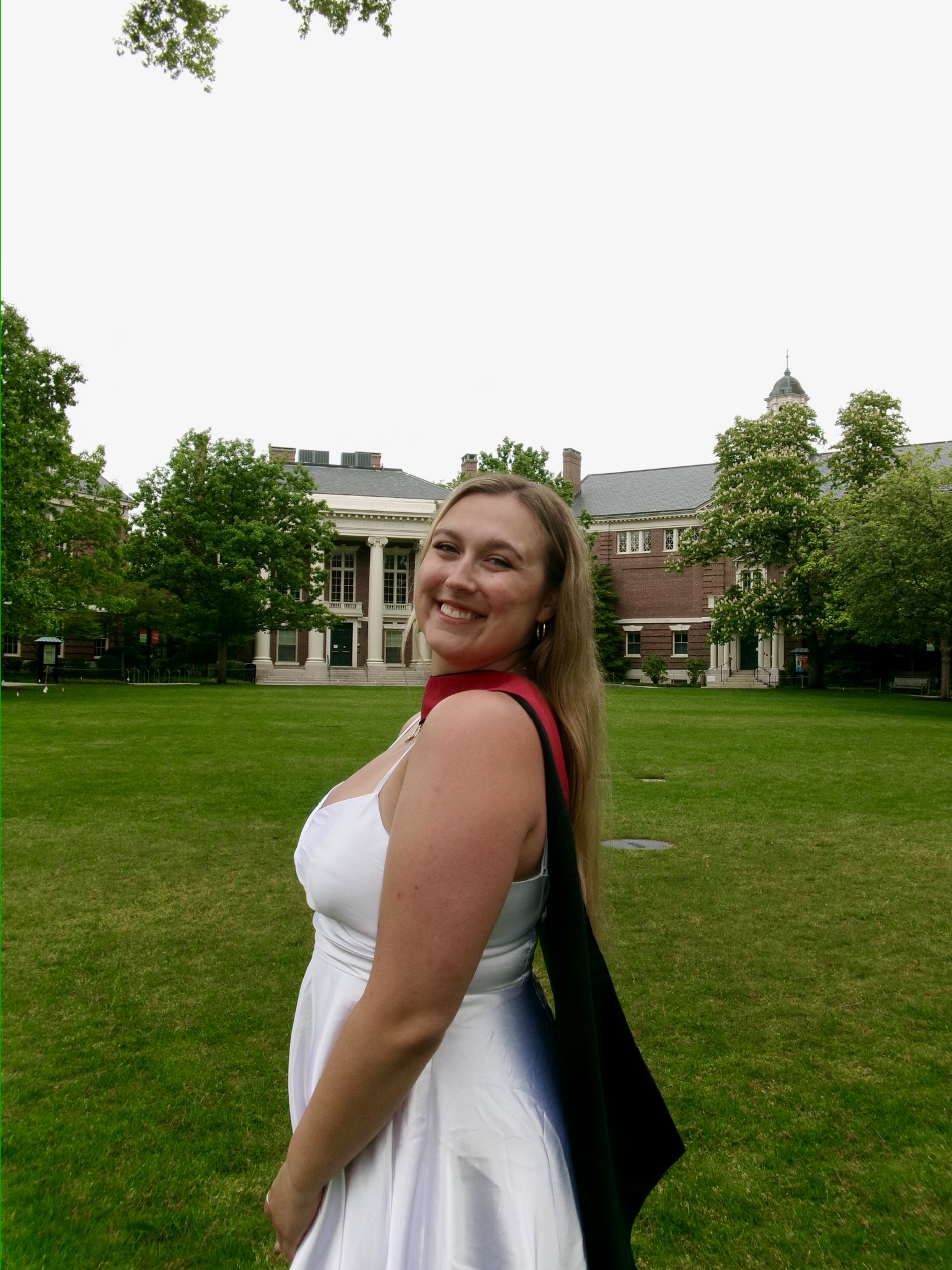 A smiling blonde woman in a white dress and red graduation stole turns toward the camera on a green university lawn, with historic brick campus buildings in the background.