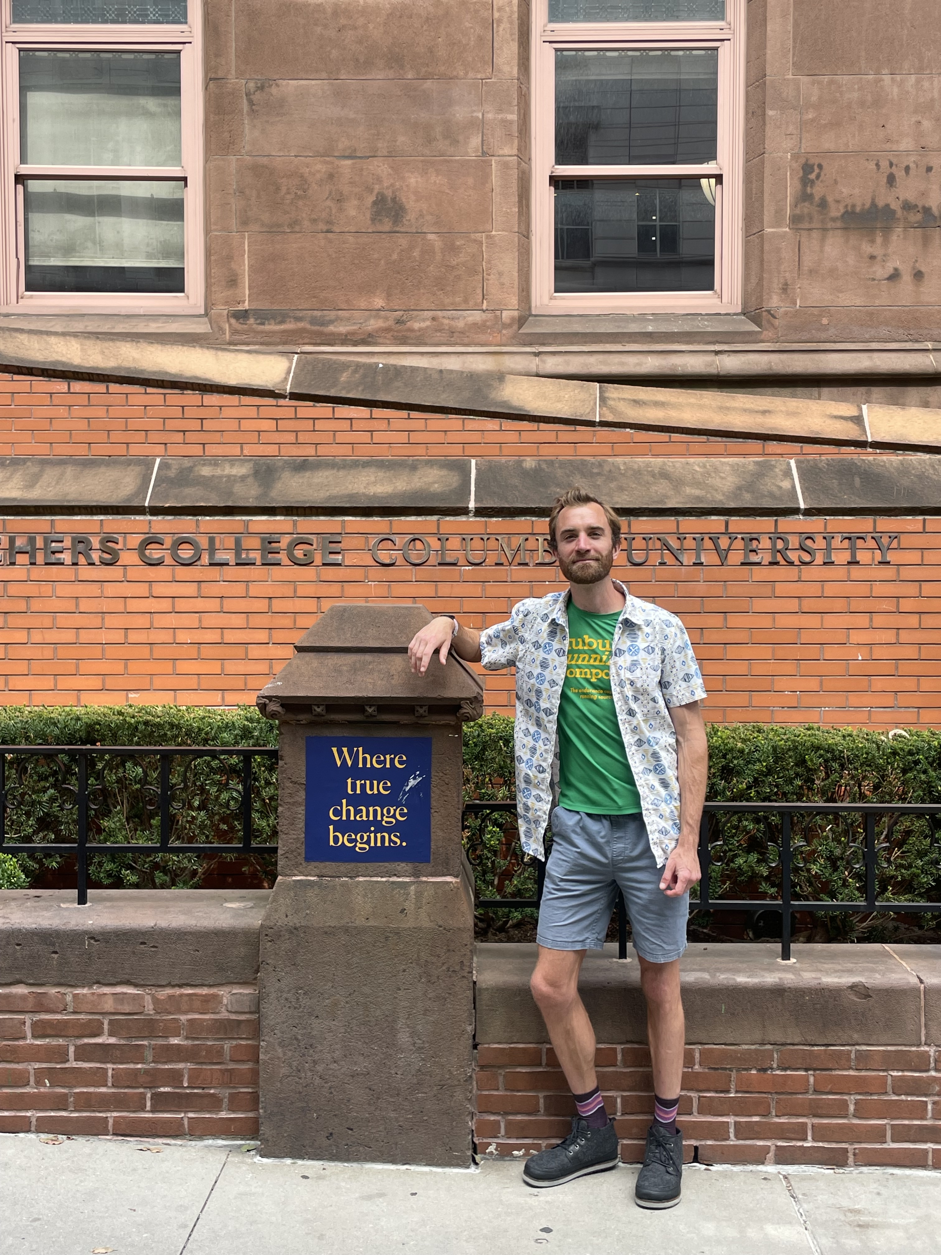 A man smiles while standing in front of the brick building for Teachers College, Columbia University. He is leaning on a pillar with a blue sign that reads, 