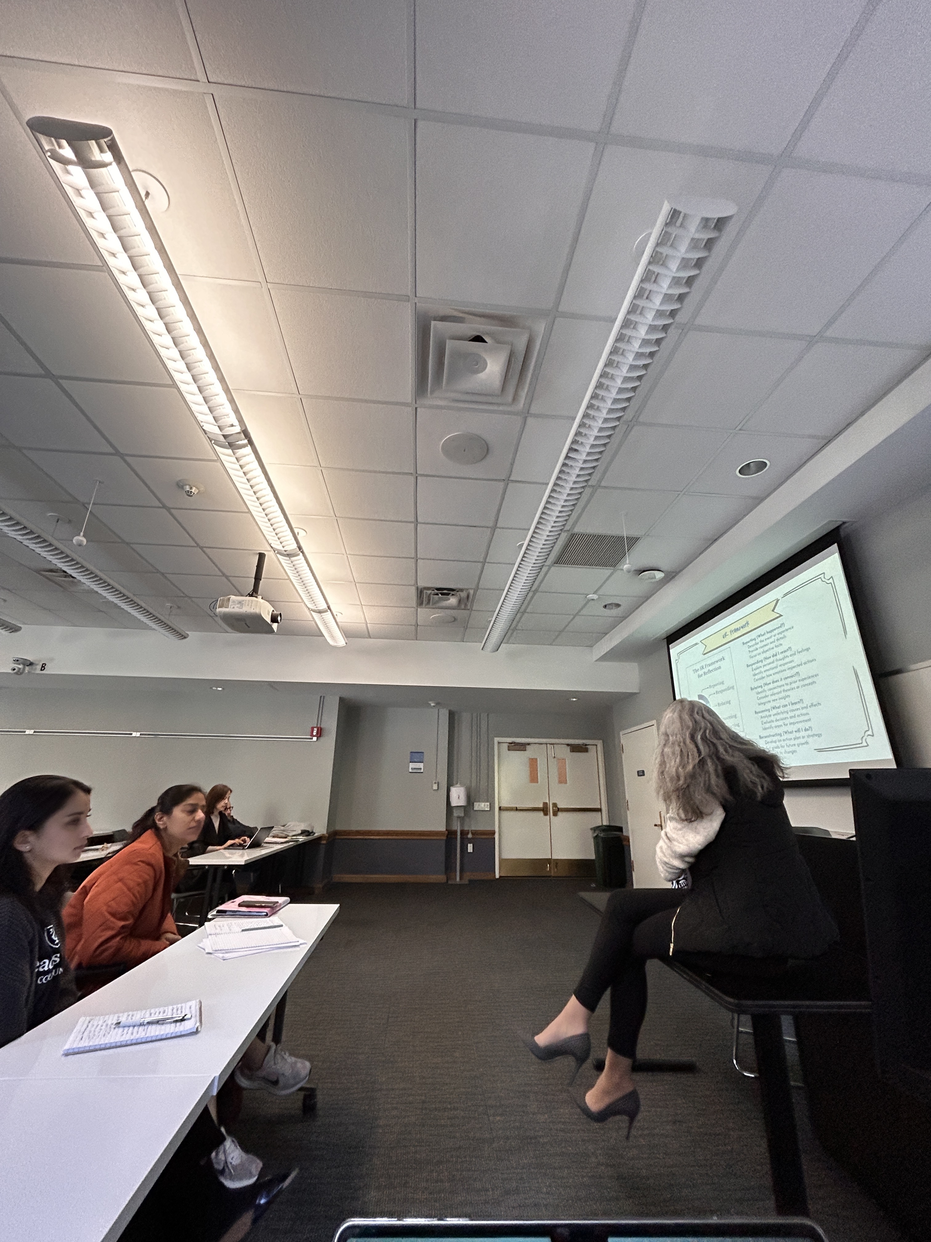 A photograph taken from behind a presenter in a classroom lecturing to a row of students. To the left of the instructor is a presentation/projection of the slide deck for reference.