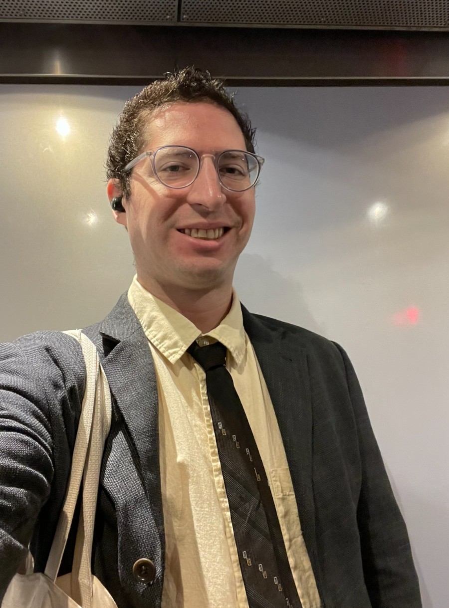 Headshot of a smiling man (Joel, he/him) dressed in a suit jacket and tie wearing glasses.