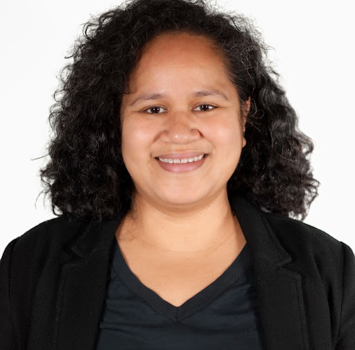 Studio headshot of Lema Moliga smiling against a plain white background. She has dark, shoulder-length curly hair and is wearing a black v-neck shirt layered under a black blazer.