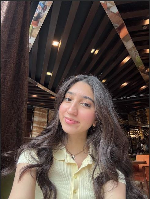 A young woman with long, wavy dark brown hair smiles gently at the camera. She is wearing a pale yellow collared shirt and a silver necklace. She is indoors, in what appears to be a restaurant with a dark, slatted ceiling.