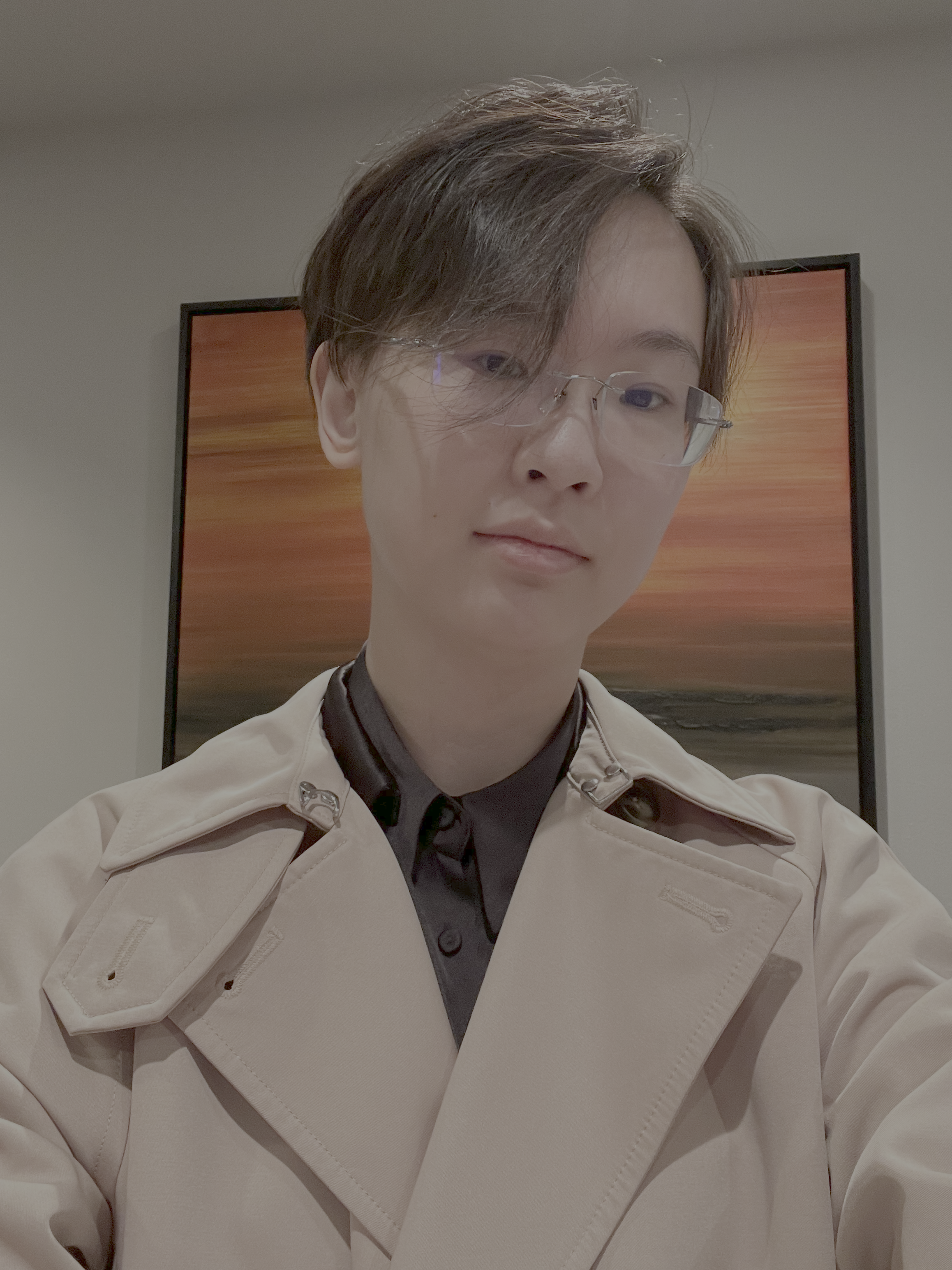 A selfie of a person with short dark hair swept to one side and frameless glasses. They are wearing a black collared shirt underneath a beige trench coat with prominent lapels. In the background, a framed artwork featuring a vivid orange, red, and yellow sunset or landscape hangs on a neutral wall.