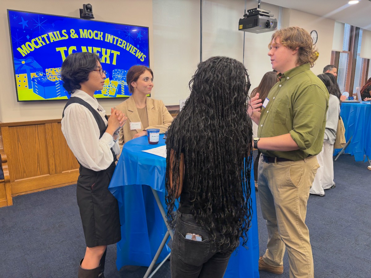 students gather around hightop table speak with alum. Screen stating 