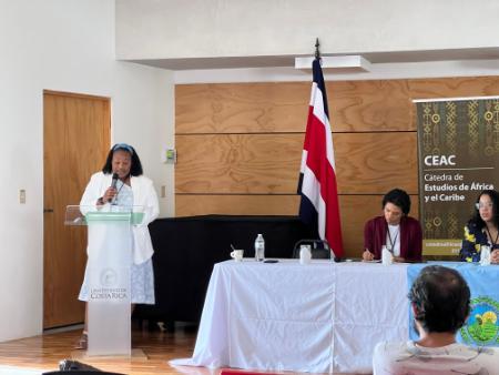 A person in a dress and white blazer is standing behind a podium, holding a microphone presenting to an audience. To the person's left is a table with seated individuals behind it with a flag and banner in the background.