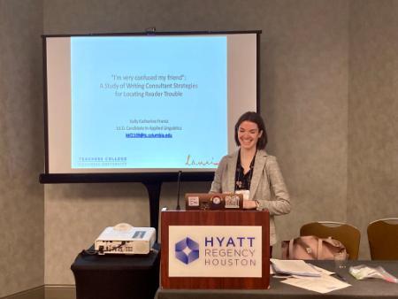 A photo of a smiling person wearing a blazer in a conference room presenting behind a podium labeled 