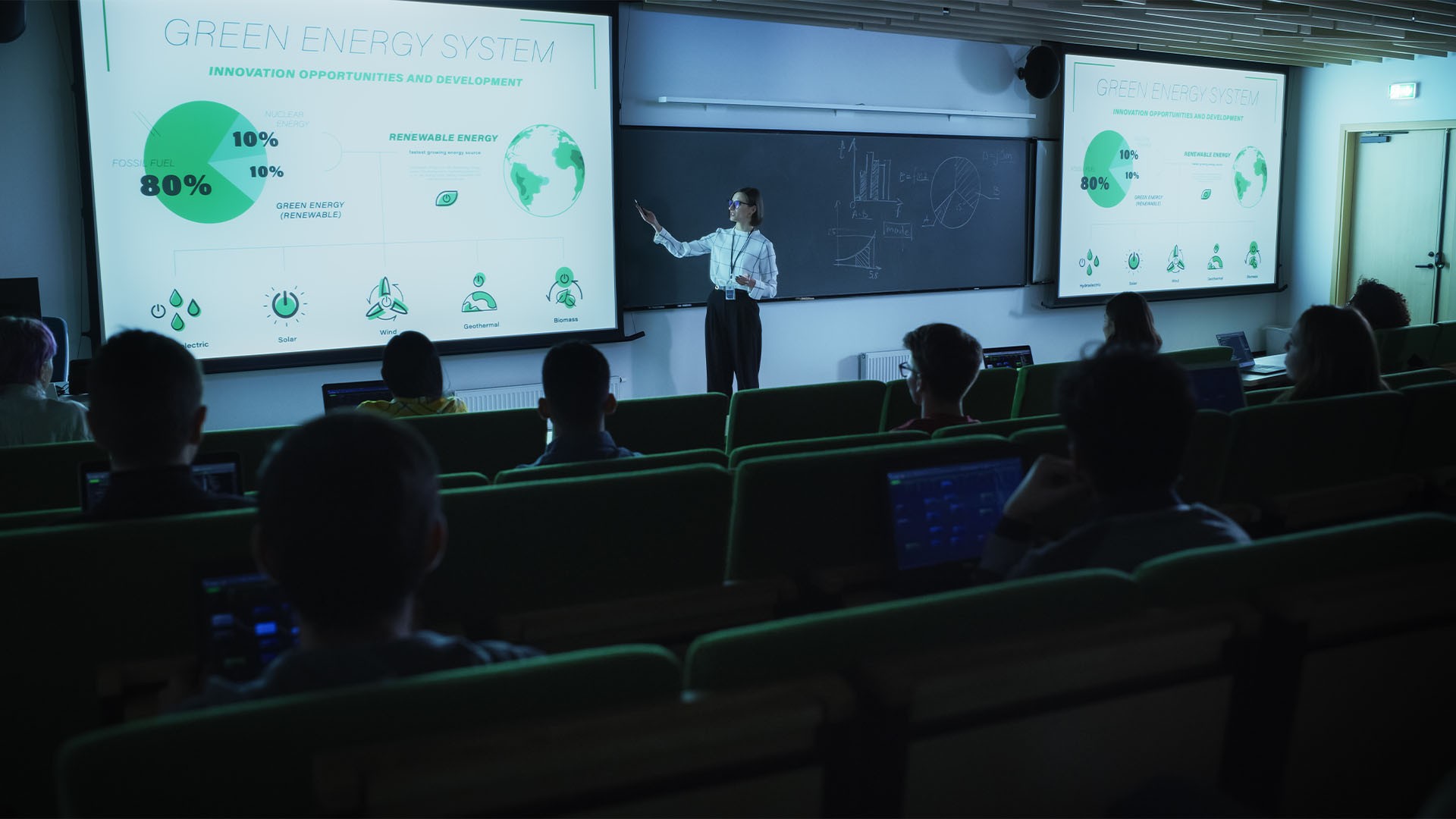 A teacher standing in front of a class of students pointing to a projector screen with information about green energy systems.