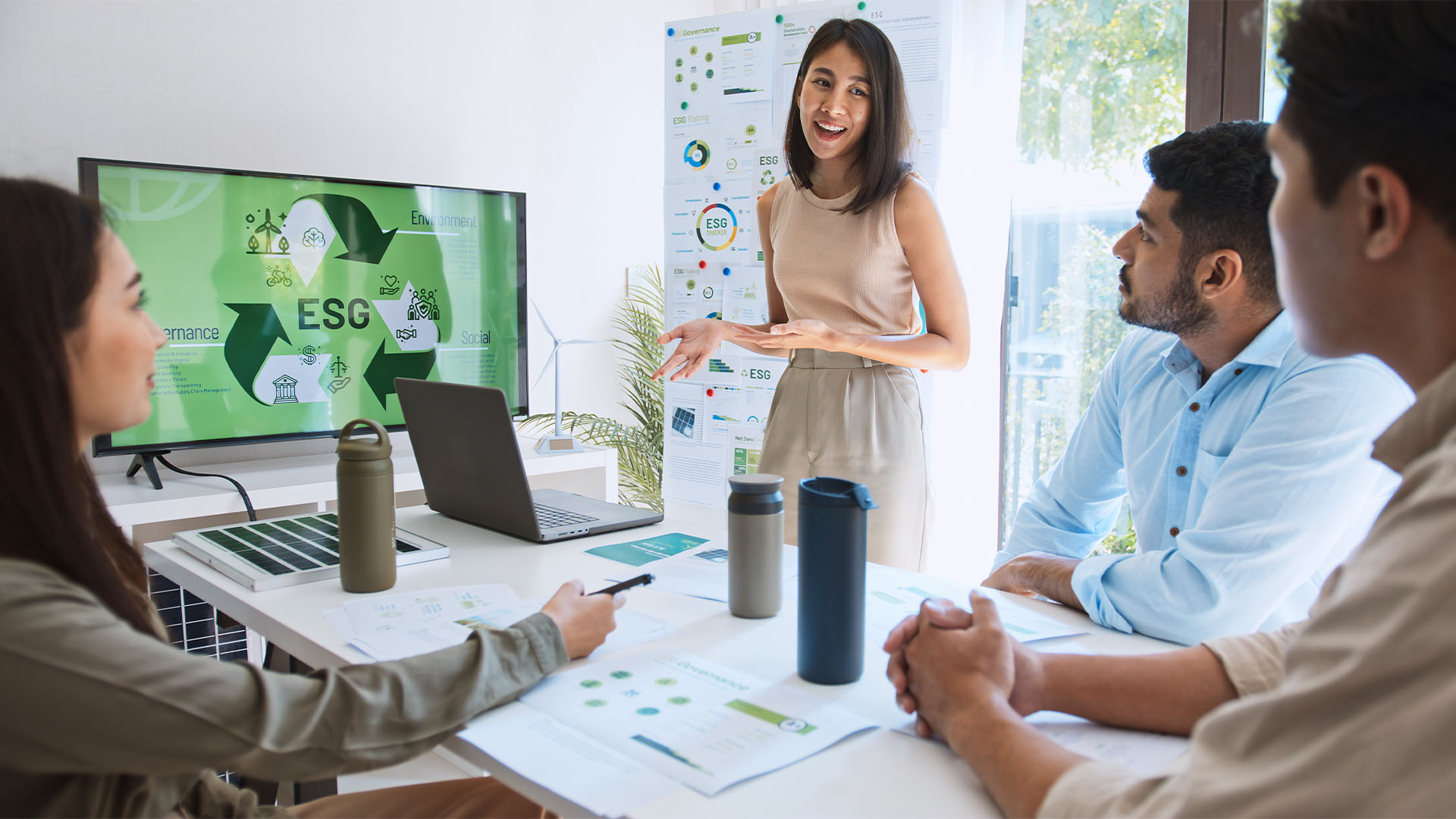 A woman giving a presentation in an office, standing in front of a screen with charts and graphs related to ESG (Environmental, Social, and Governance) topics, while three people listen.
