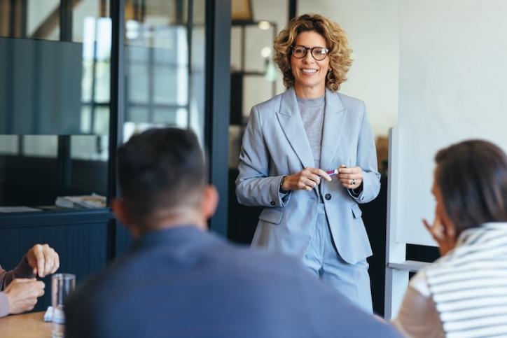 A woman in professional business attire holding a dry erase marker while standing in front of colleagues who are seated in a conference room