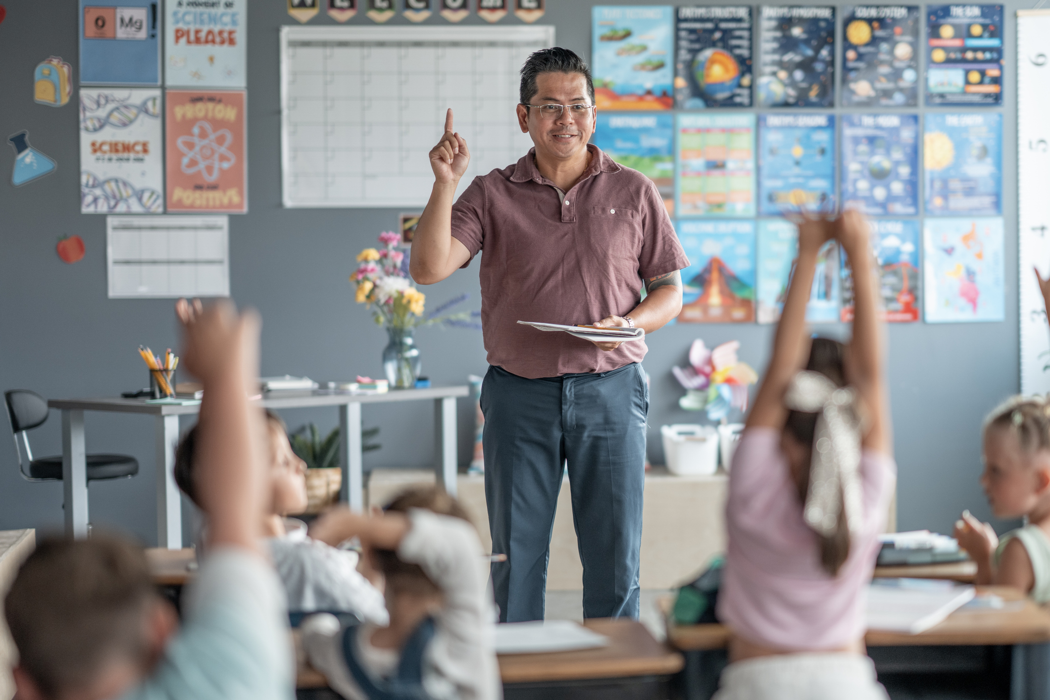 Male teacher standing in classroom in front of students raising their hands