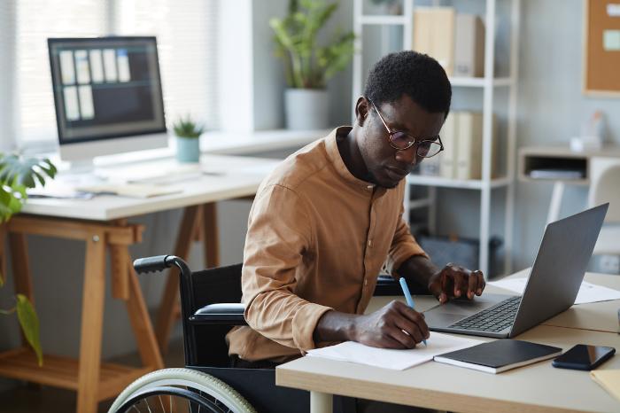 A man sitting at a desk working on a laptop and writing on a piece of paper with plants in the background