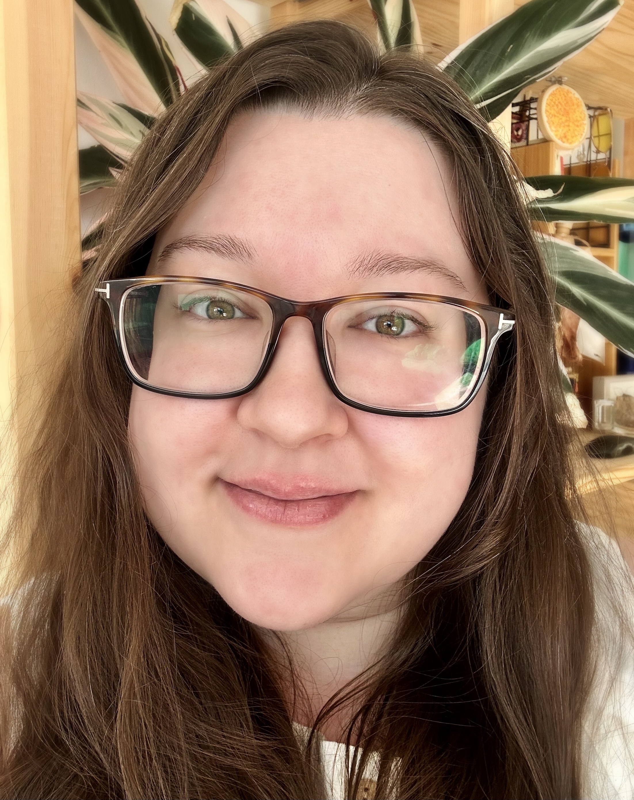 Nina Borland smiling for the camera. Nina has long brown hair, pale pinkish skin, and hazel eyes. She is wearing brown glasses.