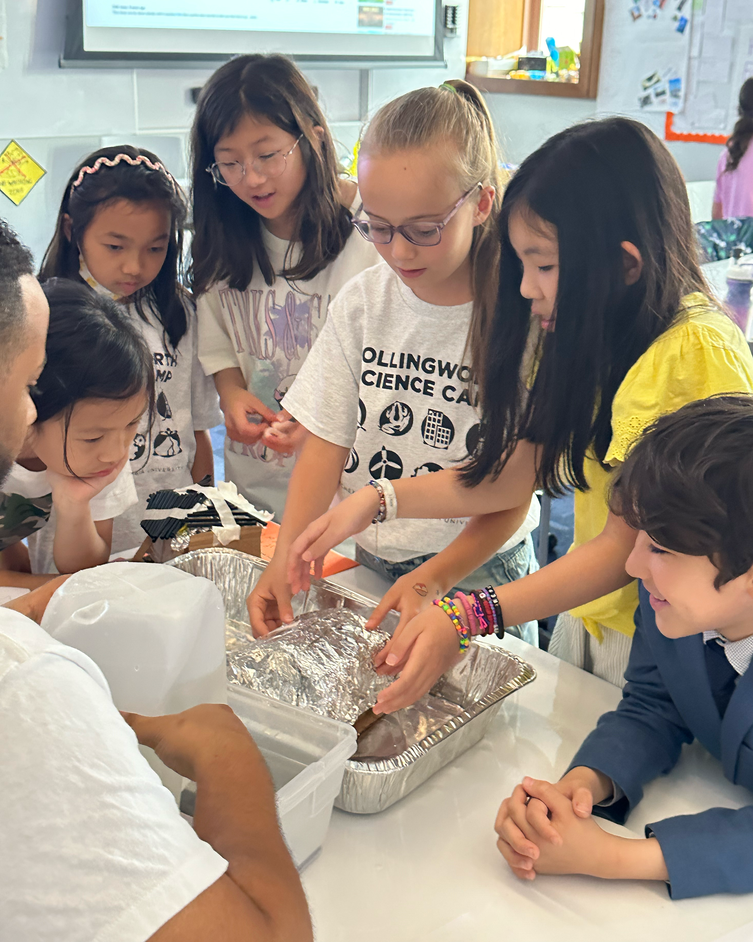 Several Children are crowed around a testing station for a protoype structure they created together. They are leaning forward and reaching out to help steady the structure.