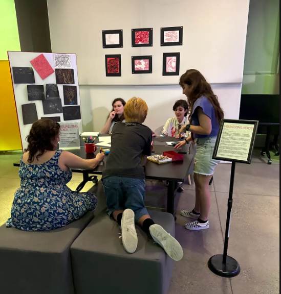 Five people gathered around a table next to a whiteboard with various printouts posted on it.