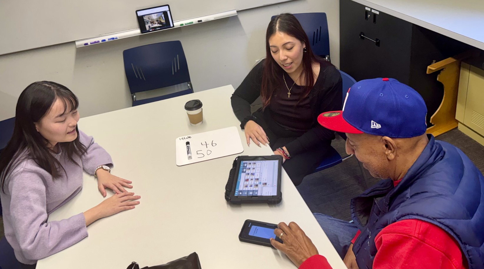 Two clinicians working with a client at a table using a tablet-based augmentative communication device, with a small whiteboard and smartphone on the table