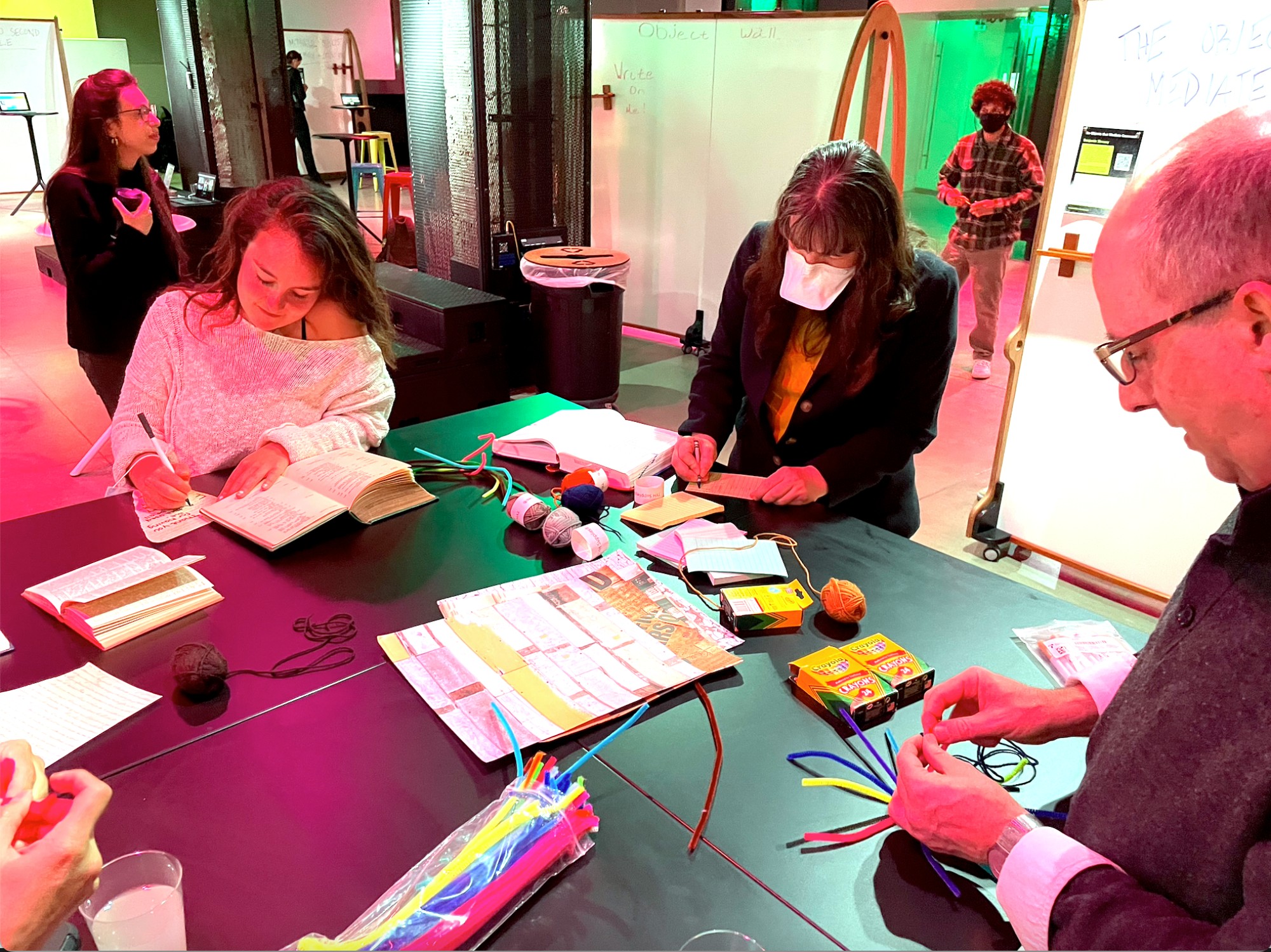 Three people stand around a table manipulating pipe cleaners, yarn, paper, and assorted craft materials. 