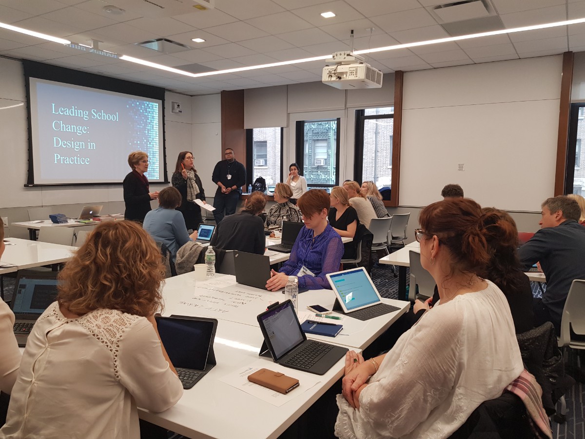 Classroom with presenters standing next to a projection screen with adult students sitting at tables with laptops