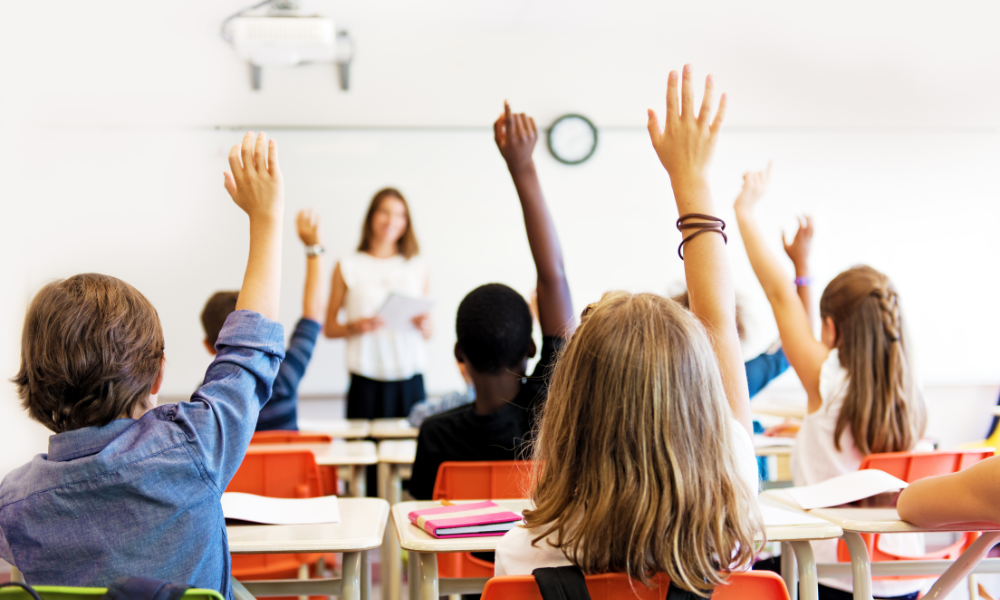 Students in a classroom raising their hands with a teacher at the front of the room
