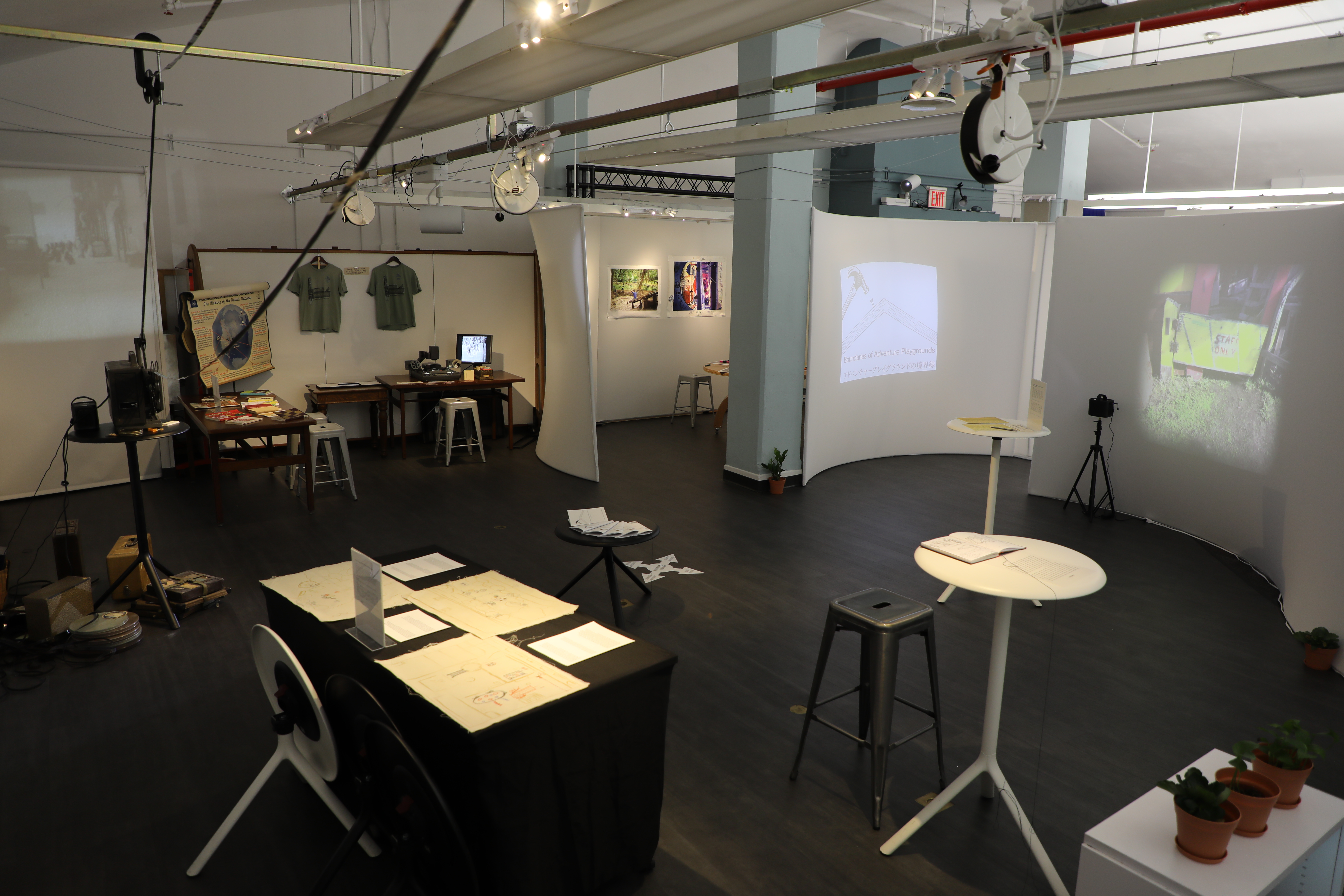 Photograph of an exhibit showing a three paper dissertation about adventure playgrounds. there are circle tables with researcher drawings, film, books, and projected images of the playgrounds on the white fabric walls.