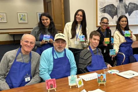 Student clinicians and participants in the Saturday Aphasia Group seated around a table displaying small painted canvases during an art activity