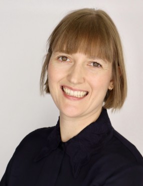 A smiling woman with short light brown hair wears a dark blouse in a professional headshot against a plain background.
