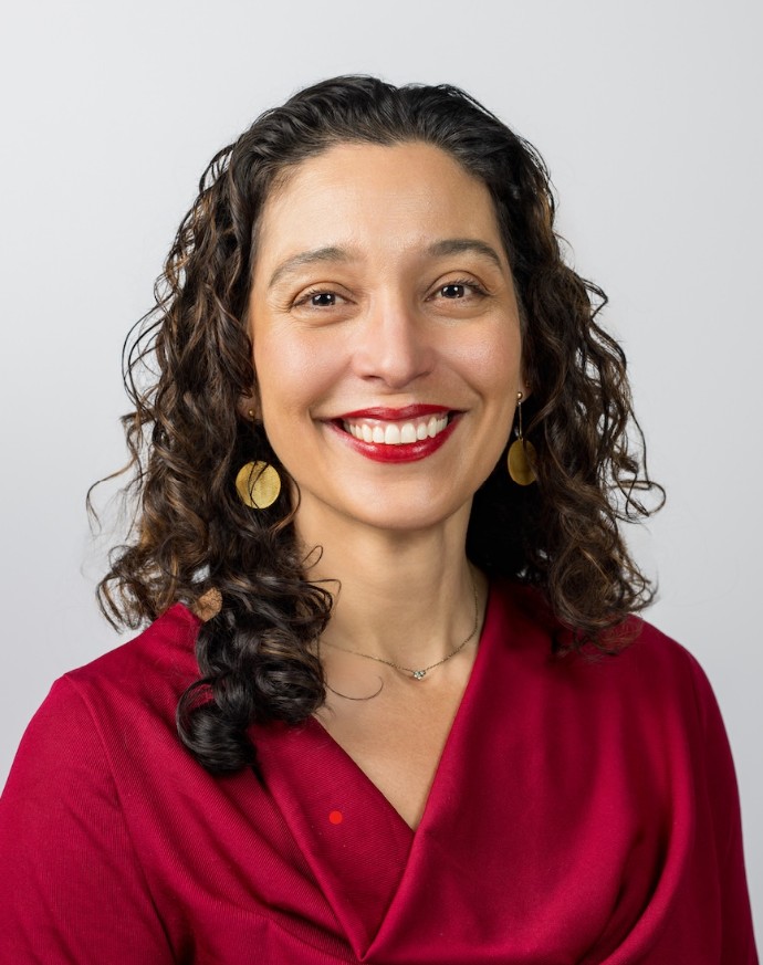 A smiling woman with curly hair wears a red top and gold earrings in a professional headshot against a plain background.