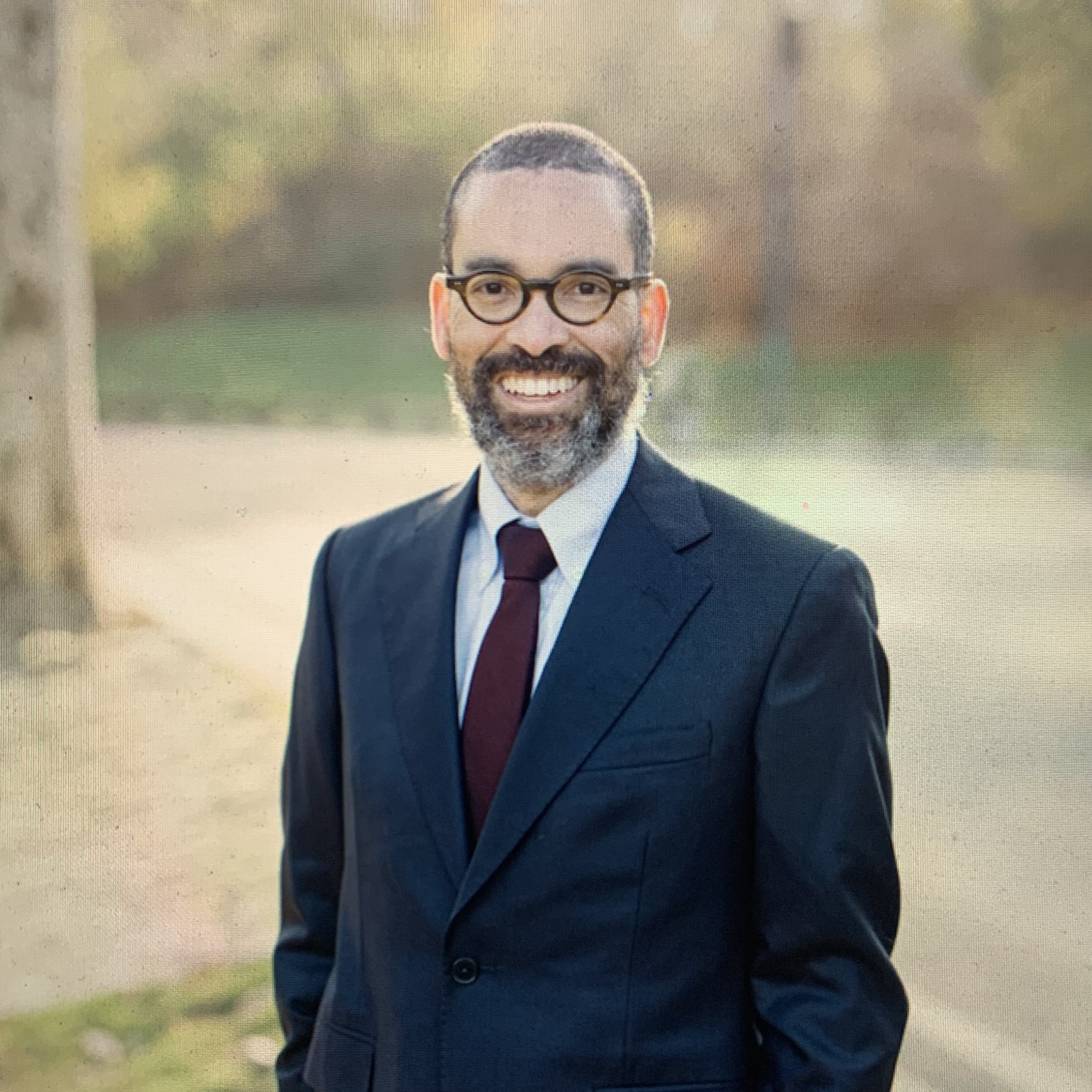 Professional portrait of a smiling man with glasses and a beard wearing a dark suit and red tie, standing outdoor