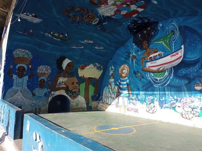 ​​Open-air soccer court with a painted wall mural surrounding it, featuring images of the ocean and Afro-Bahian individuals