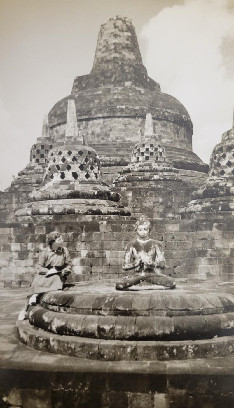 Margaret Norton (left) sits next to a statue at the Prambanan Temple complex. She gazes contemplatively at the statue to the right.