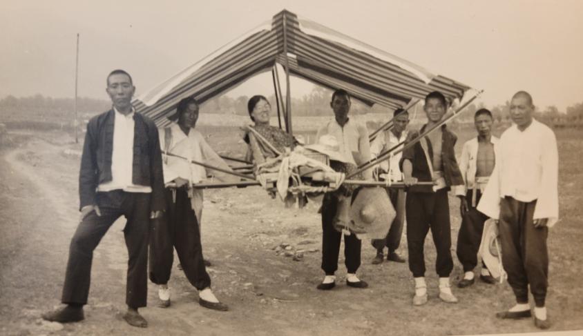Margaret Norton sits in a sedan, while numerous sedan bearers pose around her, jokingly carrying the chair.