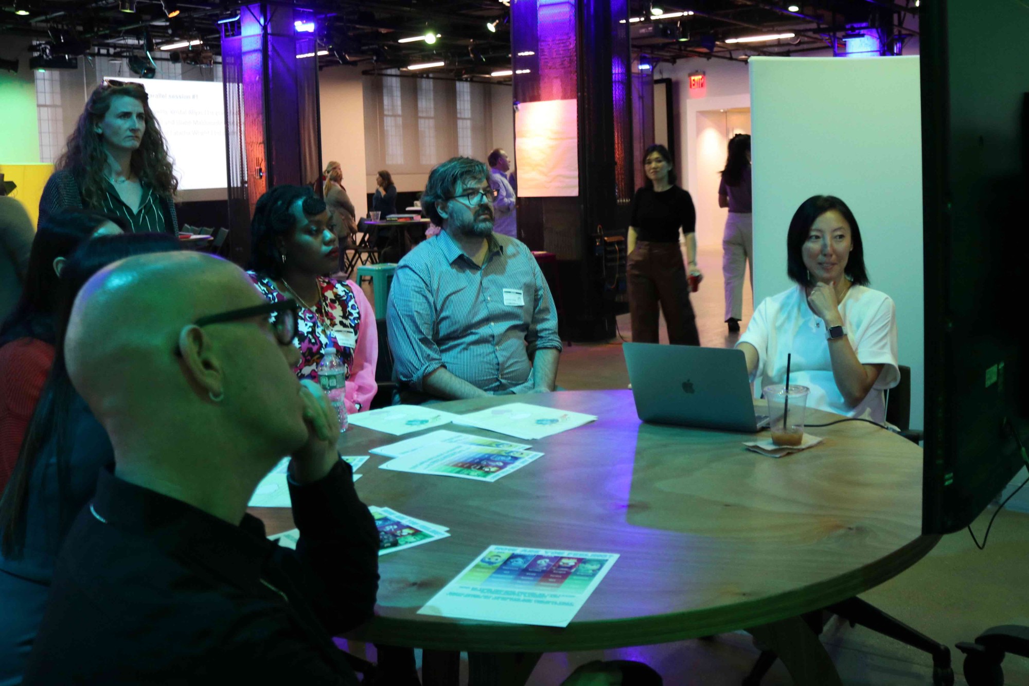 A diverse group of people sit at a round table looking at something off camera to the right. There are printed handouts on the table as well as a laptop and mostly empty iced coffee