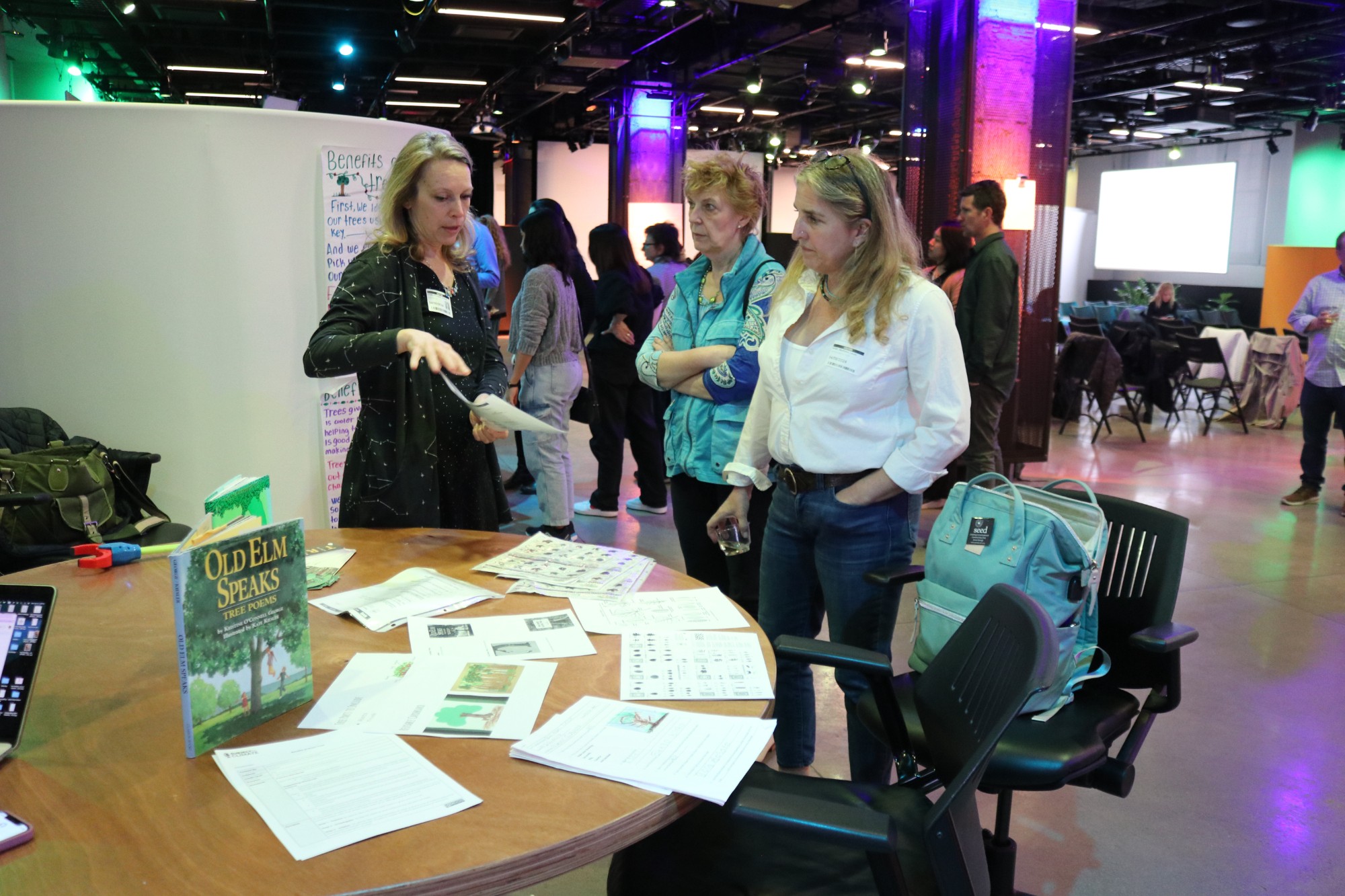 Three women stand in front of a round table that has papers and a copy 