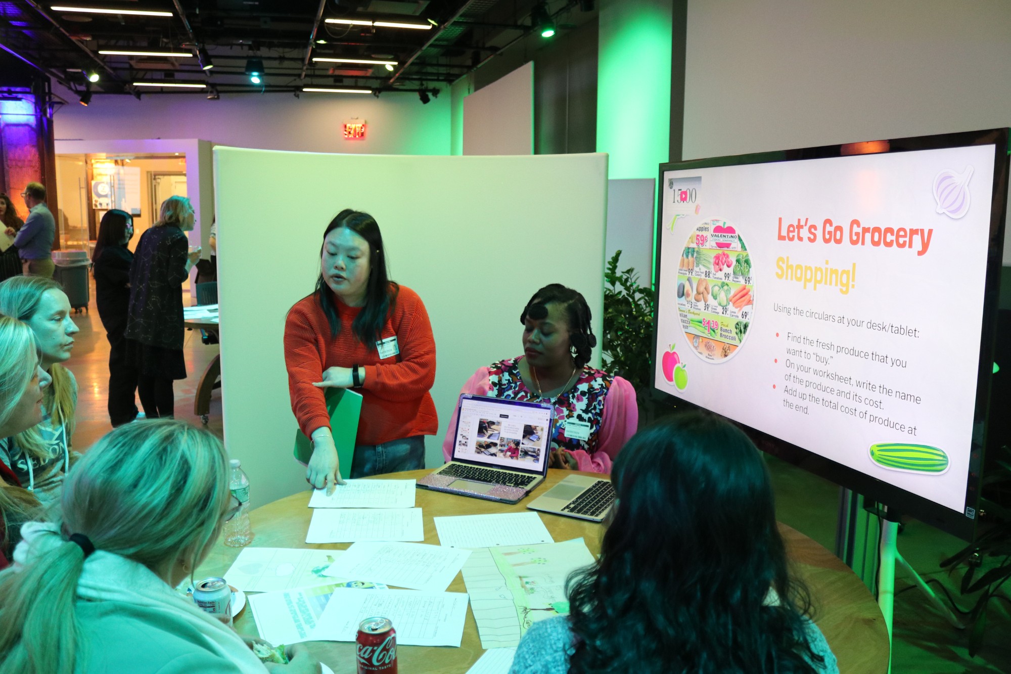 A group of four feminine-presenting people deep in discussion, sit around a round table with papers and two computers on it. On the right side of the picture there is a screen displaying a presentation slide with the header 