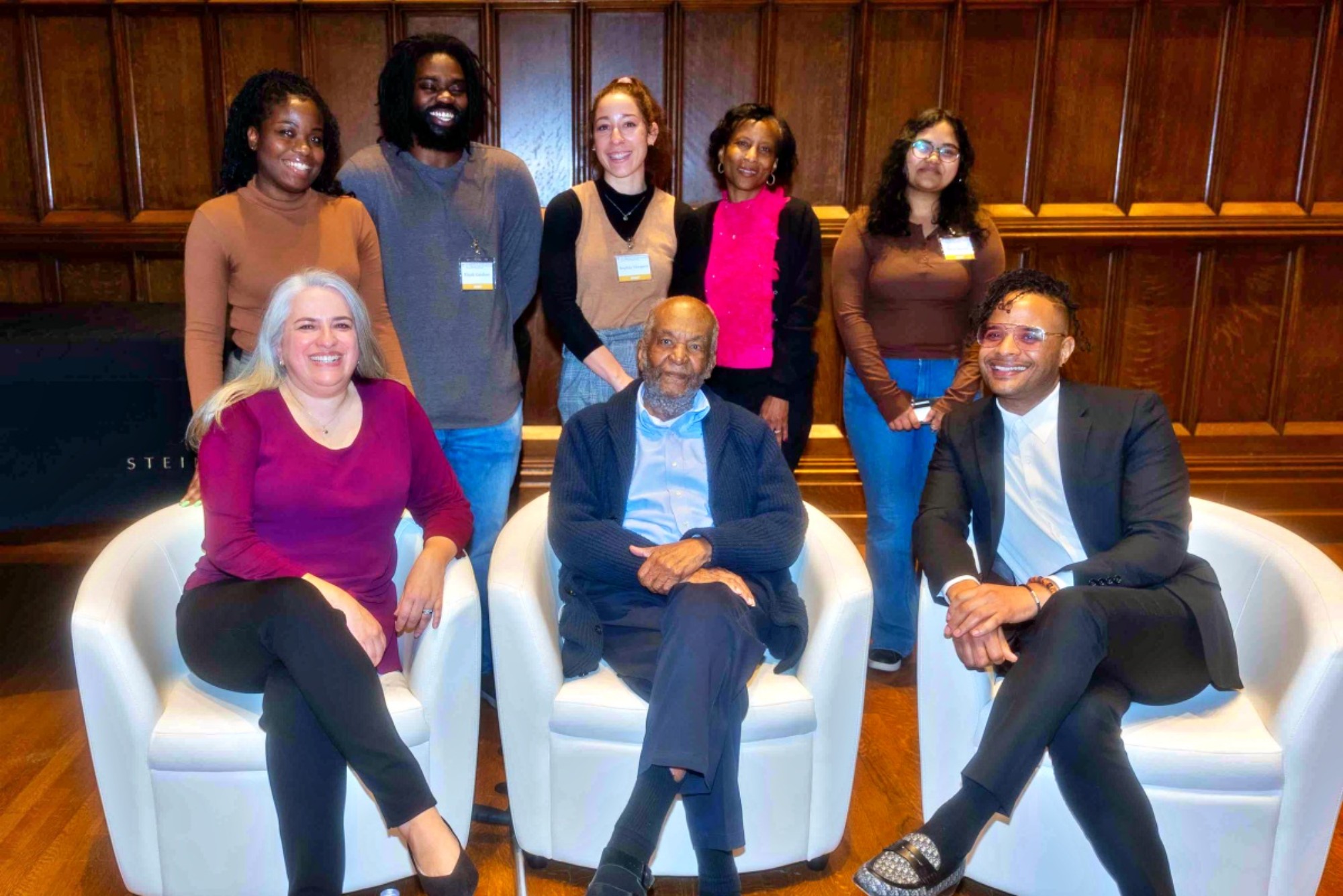 Three people, a woman and two men, sit on white chairs. Behind them are five more people. Everyone is posing for a group photo