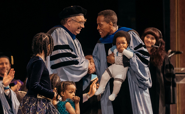 Doctoral candidate holding his son shakes hands with faculty member after being robed