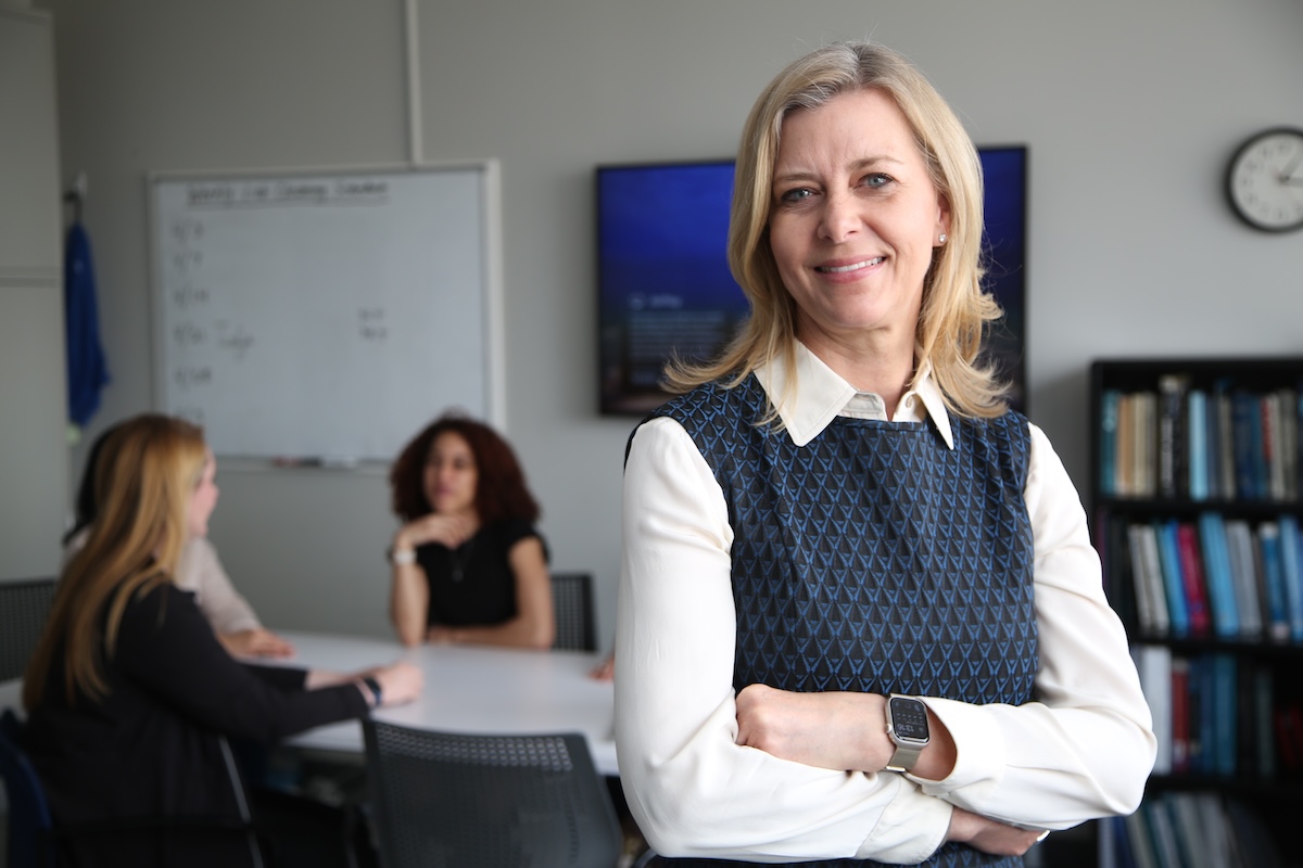 Lori Quinn, a blonde woman wearing a vest, long sleeve shirt and smart watch, smiles in a conference room with two peole in the background