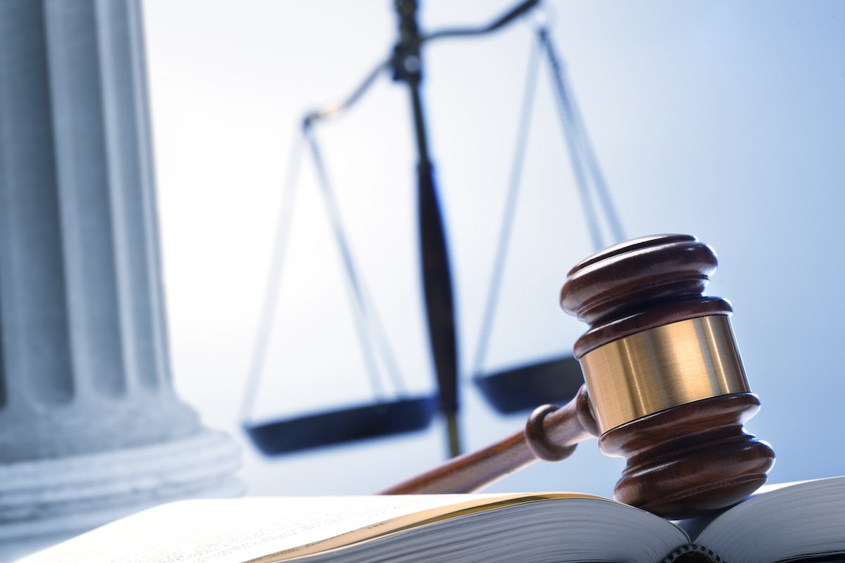 A gavel rests on top of an open law book with a justice scale and a Greek column in the background.