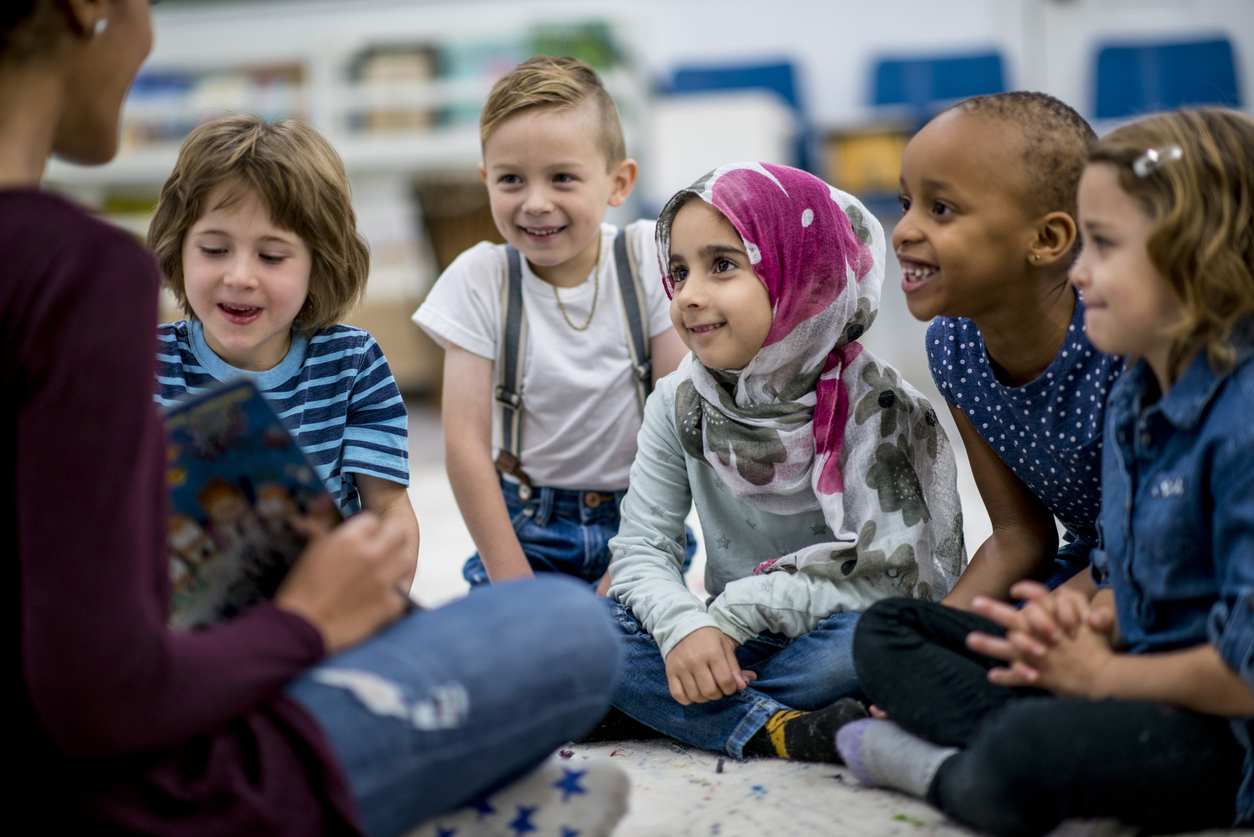 A multi-ethnic group of school children are indoors in a classroom. They are wearing casual clothing. They are sitting on the floor and eagerly listening to their teacher read a storybook.