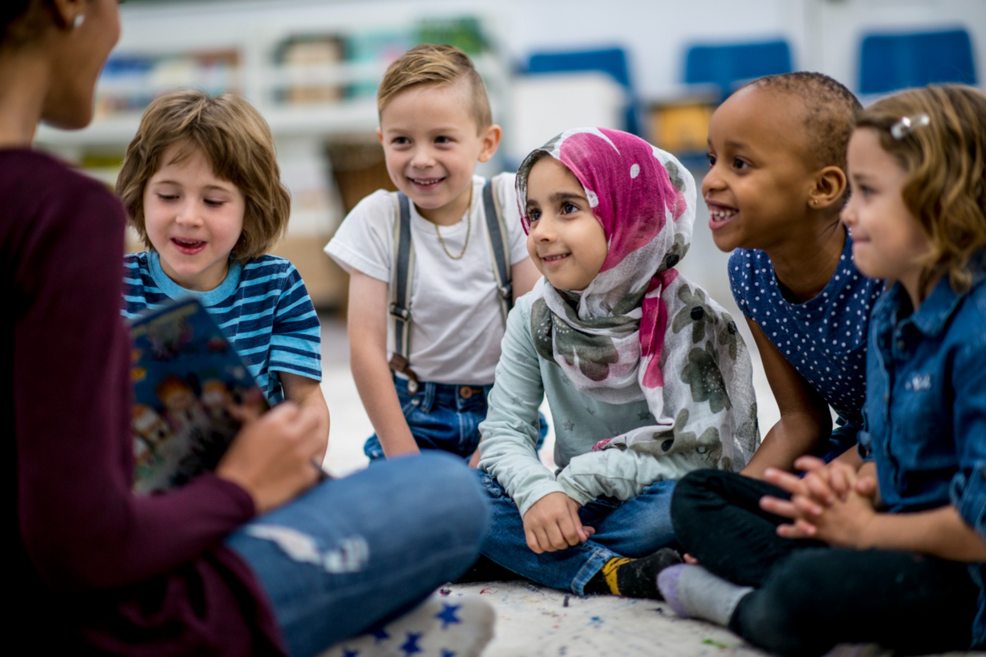 A multi-ethnic group of school children are indoors in a classroom. They are wearing casual clothing. They are sitting on the floor and eagerly listening to their teacher read a storybook.