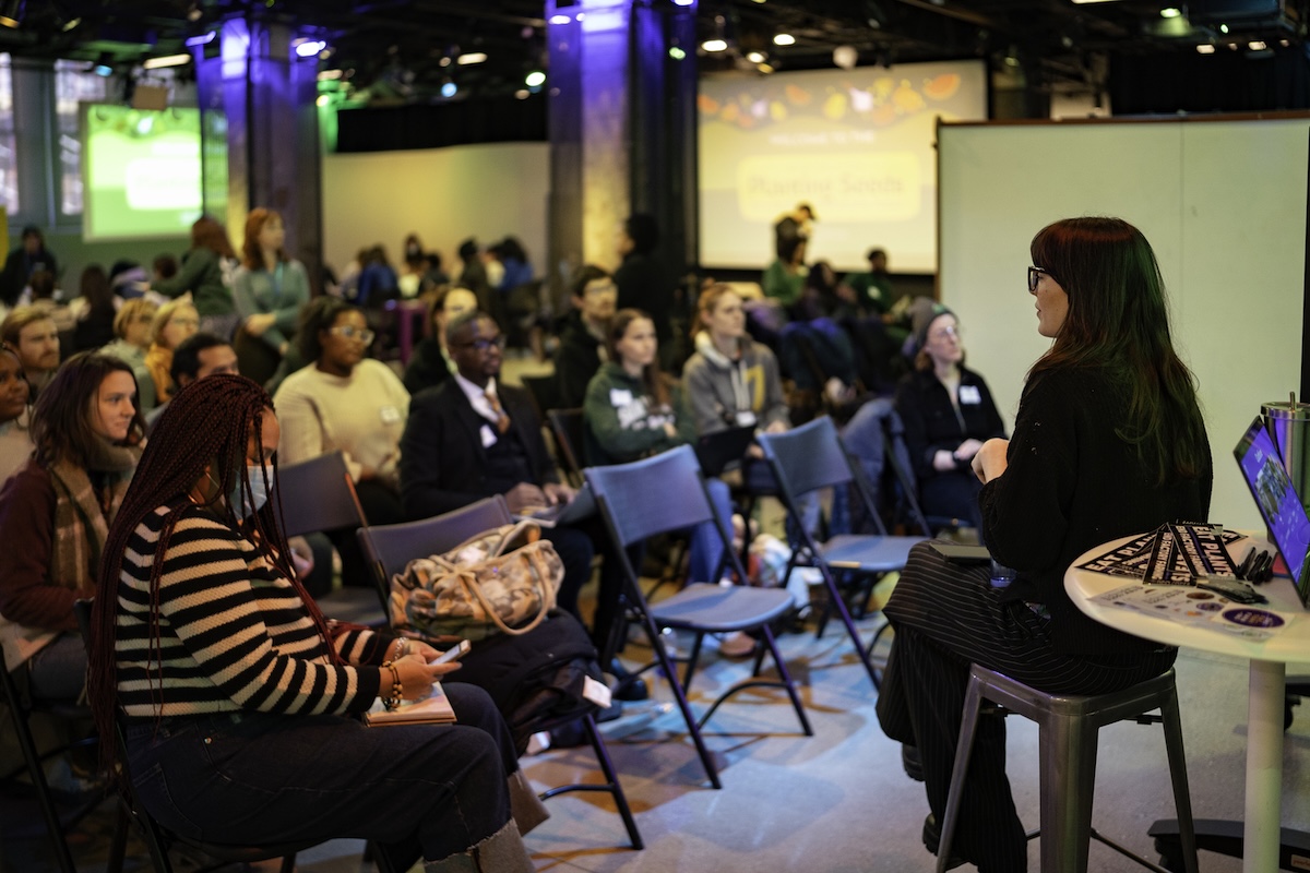 A woman, dressed in black, address a group of people sitting in chairs.