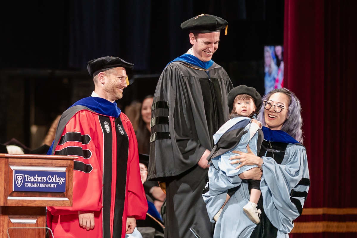 A doctoral graduate walks across the stage with her toddler, who is also dressed in a doctoral gown and hat.