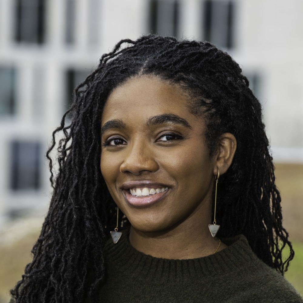 Yaa Ampofo, a woman with dreadlocks and long earrings, is smiling in front of a building