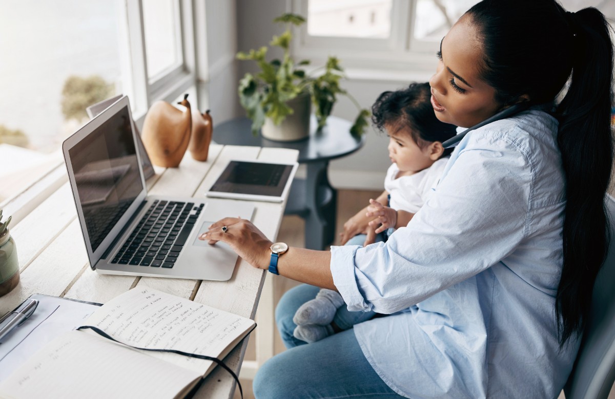 A woman holding a small child on her lap while talking on a cellphone and working on laptop